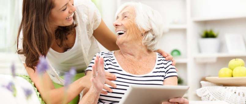 a caregiver laughing with a senior woman while she holds an ipad